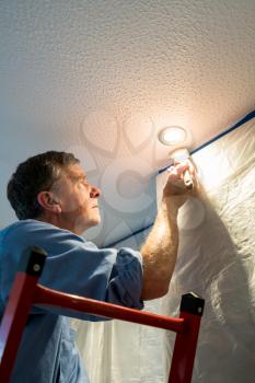 Senior adult male on ladder painting the ceiling of a room inside his home