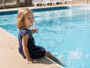 Small pre school toddler girl sitting on side of swimming pool and splashing the water with her feet