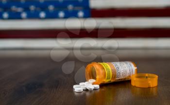 Oxycodone is the generic name for a range of opoid pain killing tablets. Prescription bottle for Oxycodone tablets and pills on wooden table with USA flag in background