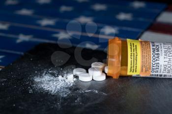 Oxycodone is the generic name for a range of opoid pain killing tablets. Prescription bottle for Oxycodone tablets and pills on slate table with USA flag in background