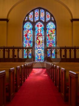 Red carpeted church aisle between pews illuminated by light from stained glass window