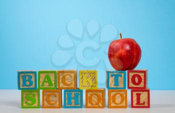 Stack of wooden blocks stacked to spell Back to School with red apple on top against blue background