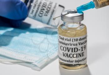 Covid-19 coronavirus vaccine with hypodermic syringe needle with droplet of liquid on tip. Gloved hand holding bottle in background