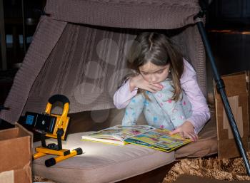 First grade girl reading a comic book with a flashlight inside home made tent with boxes and cushions
