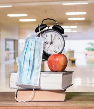 Concept for back to school with coronavirus or Covid-19 with books, alarm clock and apple with face mask against background of school corridor