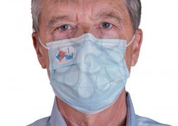I Voted sticker on senior caucasian man's face mask as he stares at the camera during the presidential election in the USA. Isolated against white