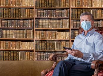 Senior caucasian man with a face mask against coronavirus reading an ebook tablet in old fashioned library of antique books