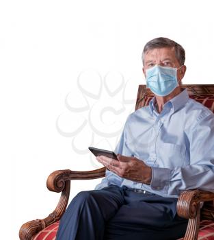 Senior caucasian adult reading from ebook in hand while seated. He is wearing a face mask against coronavirus and cutout against white