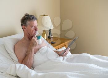 Senior retired caucasian man lying in adjustable bed on incline. He is reading and scrolling on digital tablet