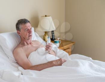 Senior retired caucasian man lying in adjustable bed on incline. He is checking his temperature for fever or signs of virus