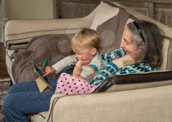 Grandmother helping small baby boy with app on a digital tablet while sitting in family room and relaxing