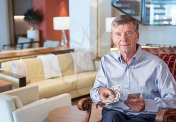 Senior caucasian man holding some US dollar bills as if handing them to the viewer. Composite into upmarket lounge of hotel or cruise ship