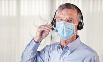 Senior caucasian man wearing facemask using headset to talk to customers or team during coronavirus epidemic