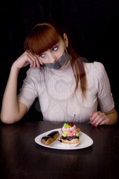 The girl, whose mouth sealed with tape sad looking at plate with cakes