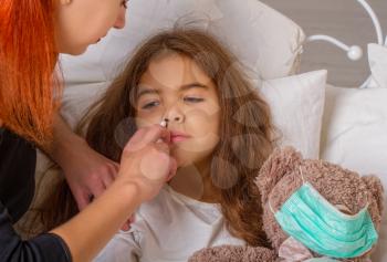 A little girl with her favorite teddy bear on whom she was wearing a gauze bandage is lying sick in bed and her mother is dripping medicine in her nose.