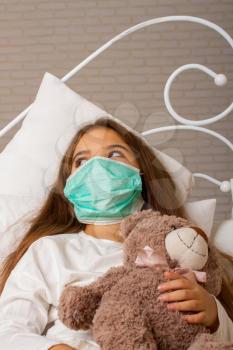 A little girl with her favorite toy bear is lying sick in bed with a gauze bandage over her face.