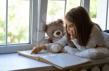 Little Girl lies on the windowsill next to her favorite toy bear cub and reads an old big book