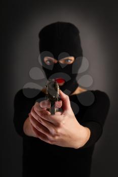 Slim sexy girl in dark inconspicuous clothes hiding her face under a balaclava hat with a gun on a gray background