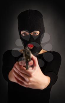Slim sexy girl in dark inconspicuous clothes hiding her face under a balaclava hat with a gun on a gray background