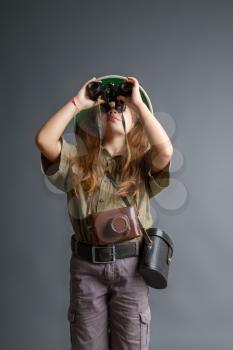 a little girl in a tropical khaki uniform and a cork helmet looks through binoculars somewhere upwards