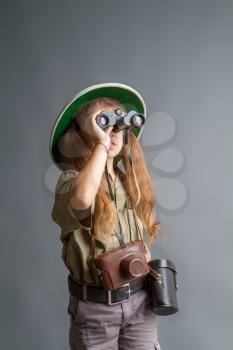 a little girl in a tropical khaki uniform and a cork helmet looks through binoculars somewhere upwards