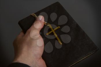 a man's hand holds a bible in a dark binder a metal cross on a chain close-up