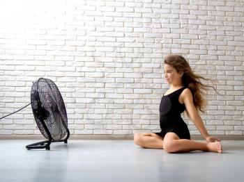 little girl struggling with heat and sitting in front of a fan with flying hair in an apartment on a bricky light wall background