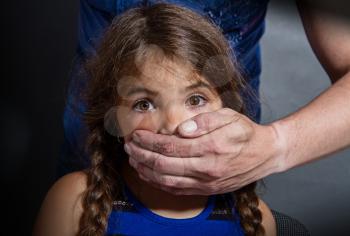 the rough hand of an adult man closes the palm of the mouth of a little girl with frightened eyes against a dark wall
