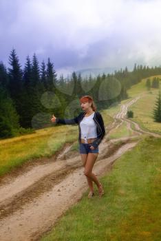 the girl stands on a rain-soaked mountain dirt road stretching out her hand and raising her finger for auto-stop