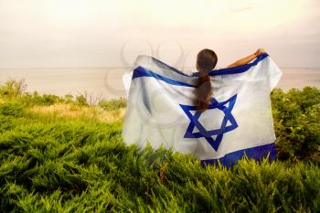 a little girl with an Israeli flag raised in her hands and behind her stands on the shore of a pond in a thicket of grass.