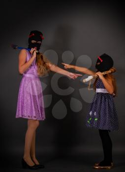 Two hooligans of a teenager girl in bright elegant dresses, but in gangster hats, balaclava with heavy clubs, laugh at each other because of their clothes on a dark background