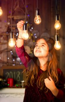 little girl is played with old lamps decorating a room against the background of a wooden wall and a Christmas tree made of branches