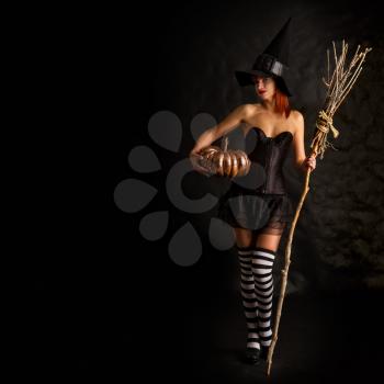 sexy girl dressed as a medieval witch stands with a broom and pumpkin in hand