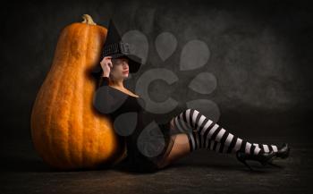 tired girl in halloween witch costume with hat and striped stockings rests sitting leaning with his back to a big pumpkin