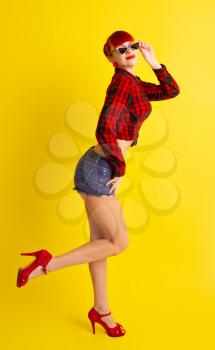 Red-haired girl dressed in retro style in a red checkered shirt and shorts and sunglasses posing on a bright yellow background