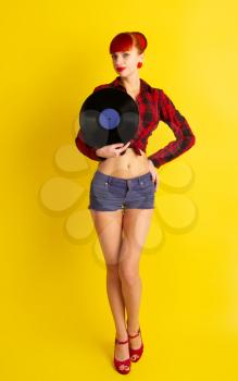 Red-haired girl dressed in retro style in a red checkered shirt and shorts on keeps a vinyl record on a yellow background
