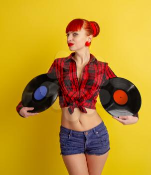 Red-haired girl dressed in retro style in a red checkered shirt and shorts on keeps a vinyl record on a yellow background