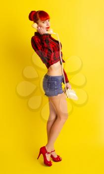 Pretty girl in knotted plaid shirt and shorts with retro phone talking to someone on a bright yellow background