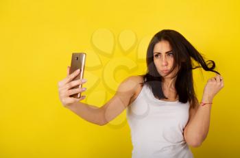Young cute girl in casual clothes dabbles and grimaces and takes a selfie on her smartphone against a bright yellow background