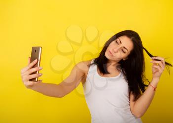 Young cute girl in casual clothes dabbles and grimaces and takes a selfie on her smartphone against a bright yellow background