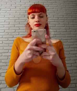 Young red-haired girl is reading something thoughtfully on her smartphone against a gray brick wall
