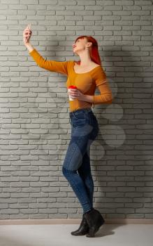 Young red-haired girl with a cup of coffee makes selfie with her smartphone on a background of gray brick wall