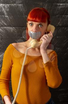 Conceptual picture. Young red-haired girl tries to talk on the phone with sealed with adhesive tape with her lips so that she keeps silence