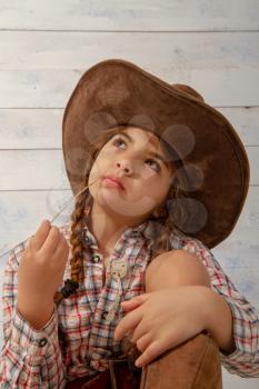 A little girl in a wide-brimmed cowboy hat wearing a traditional dress and high boots is eating a straw on a light wooden background