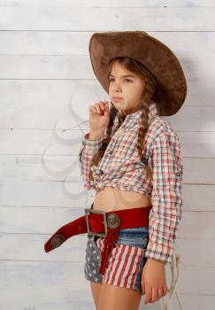 A little girl in a wide-brimmed cowboy hat wearing a traditional dress and with a lasso is eating a straw on a light wooden background.