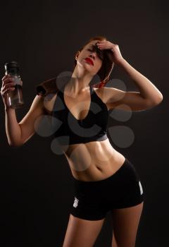 Young tired girl in short sportswear and towel on her shoulders drinks water from a special bottle after fitness classes
