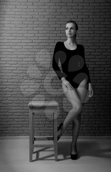 Young sensual girl in a black swimsuit and fishnet stockings and high heels with a high gray stool against a dark brick wall Black and white.