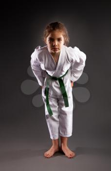 A little girl in a traditional white kimono for karate and a green belt performs training exercises against a dark background.