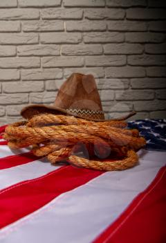 Traditional cowboy brown hat, lasso of coarse rope and a US flag against a dark brick wall