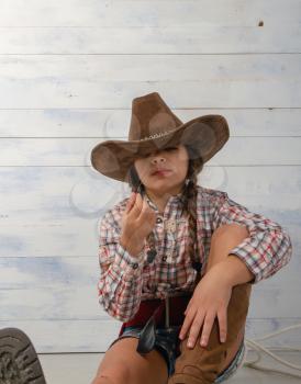 A little girl in a wide-brimmed cowboy hat wearing a traditional dress and high boots with a lasso is eating a straw on a light wooden background.